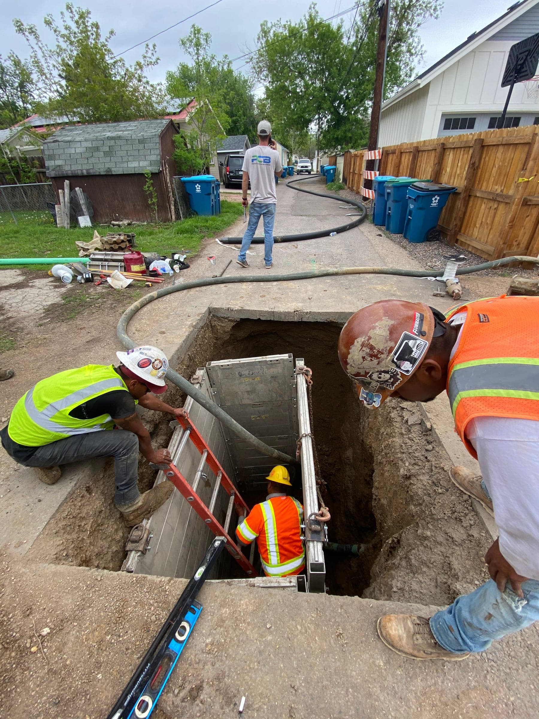 Crew lowering into a trench box in a residential alley, one worker on the ladder and two watching from the edge