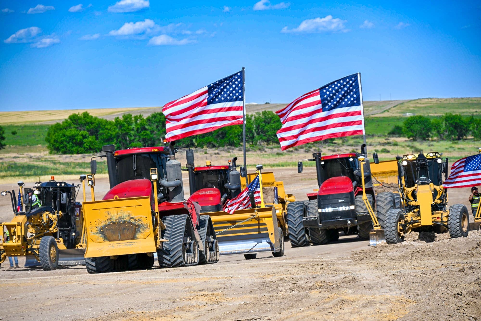 Red tractors, yellow scrapers, and graders lined up with American flags mounted on the rigs