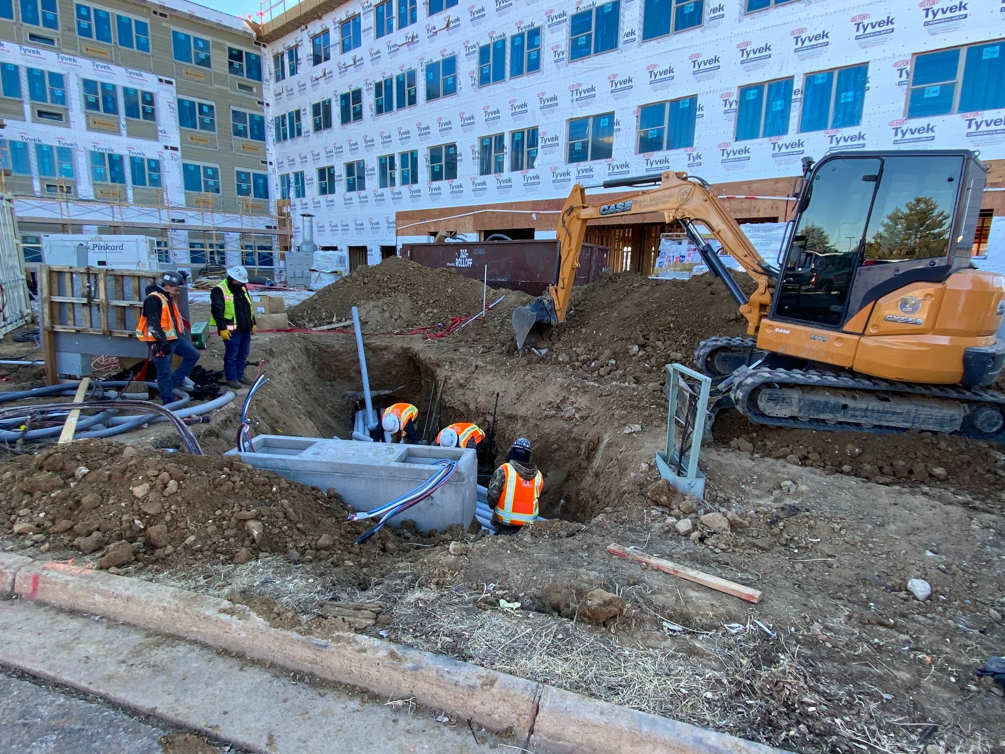 Excavator and crew working an electrical trench beside a new building