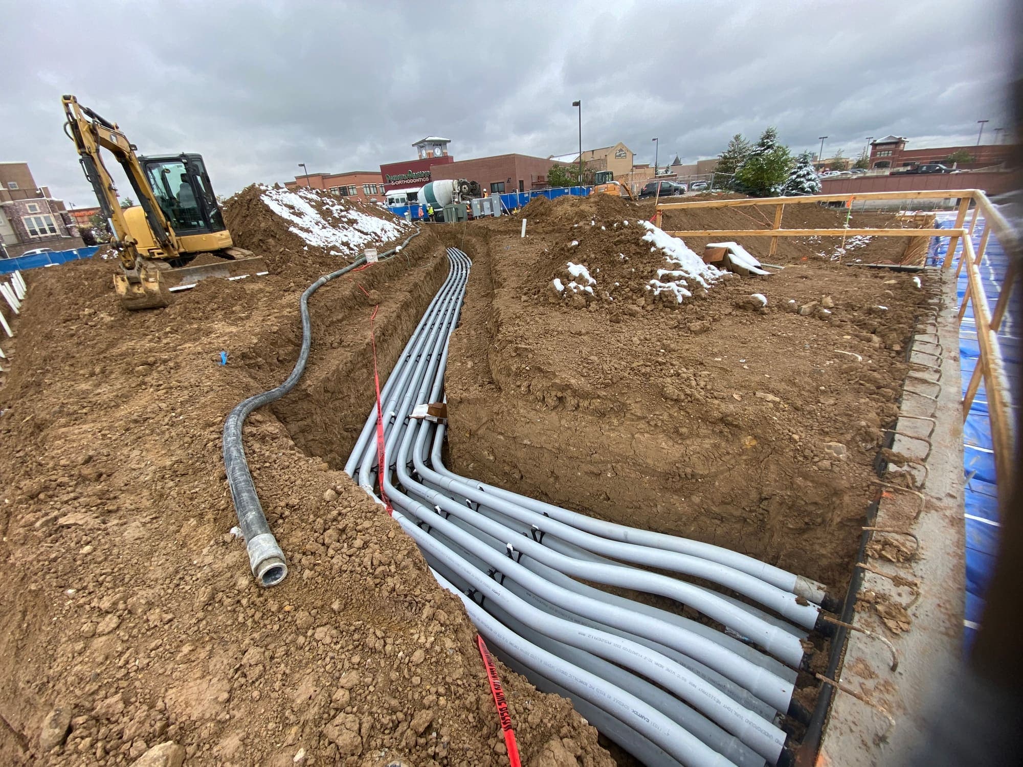 Multiple gray conduits sweeping through a trench with an excavator behind