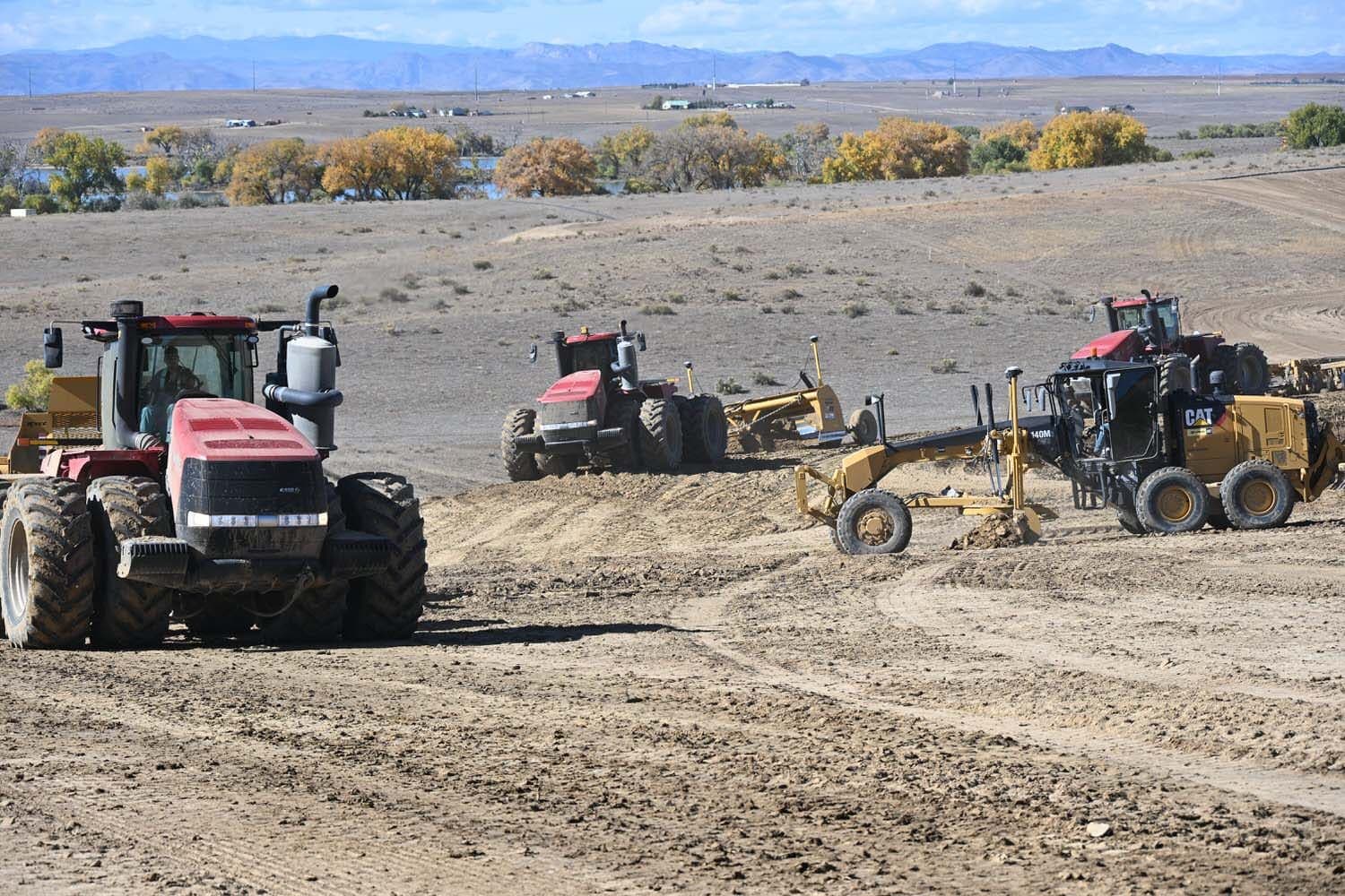 Red tractors and a motor grader working a graded site near a line of trees