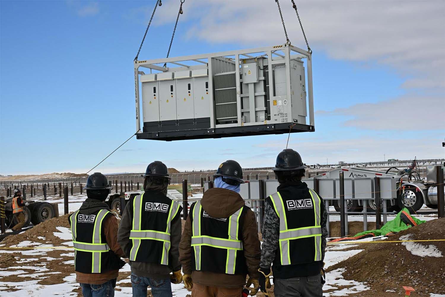 Four BME crew members in safety vests watching a crane set an equipment enclosure on a solar site