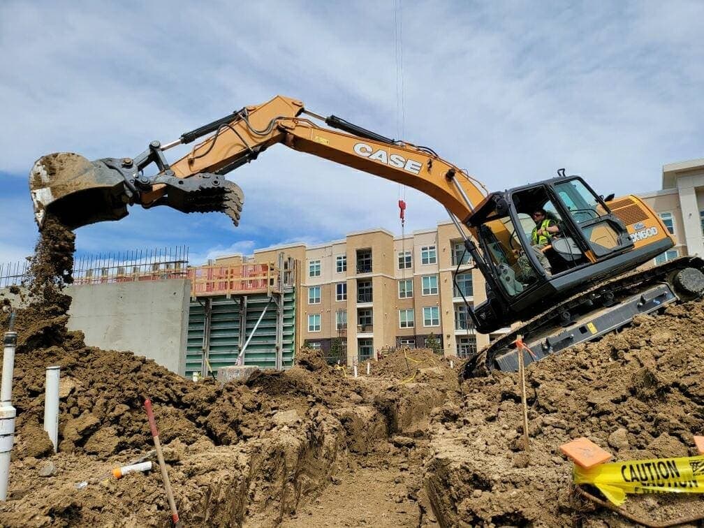 An excavator lifting a bucket of dirt in front of a new apartment building under construction