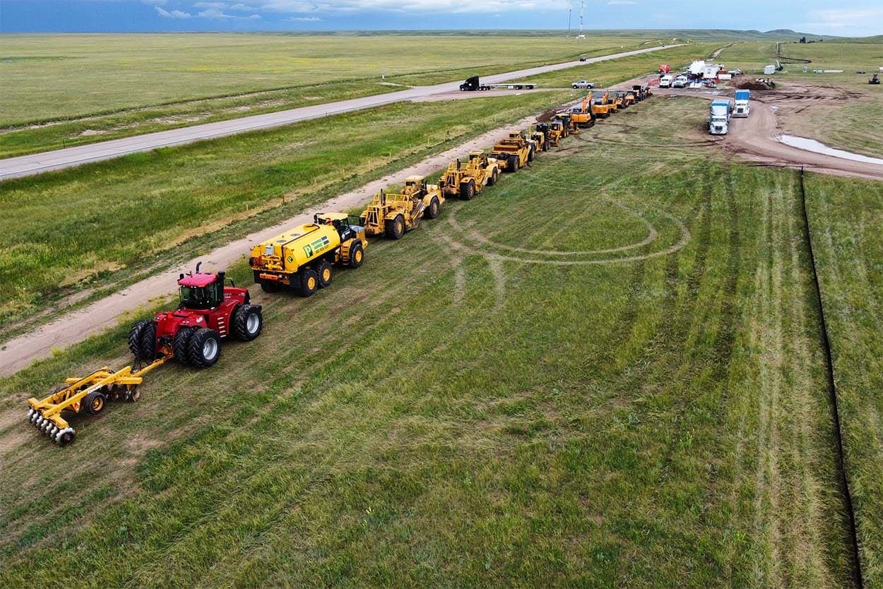 Drone view of a long line of tractors, scrapers, and a water truck along a grass road