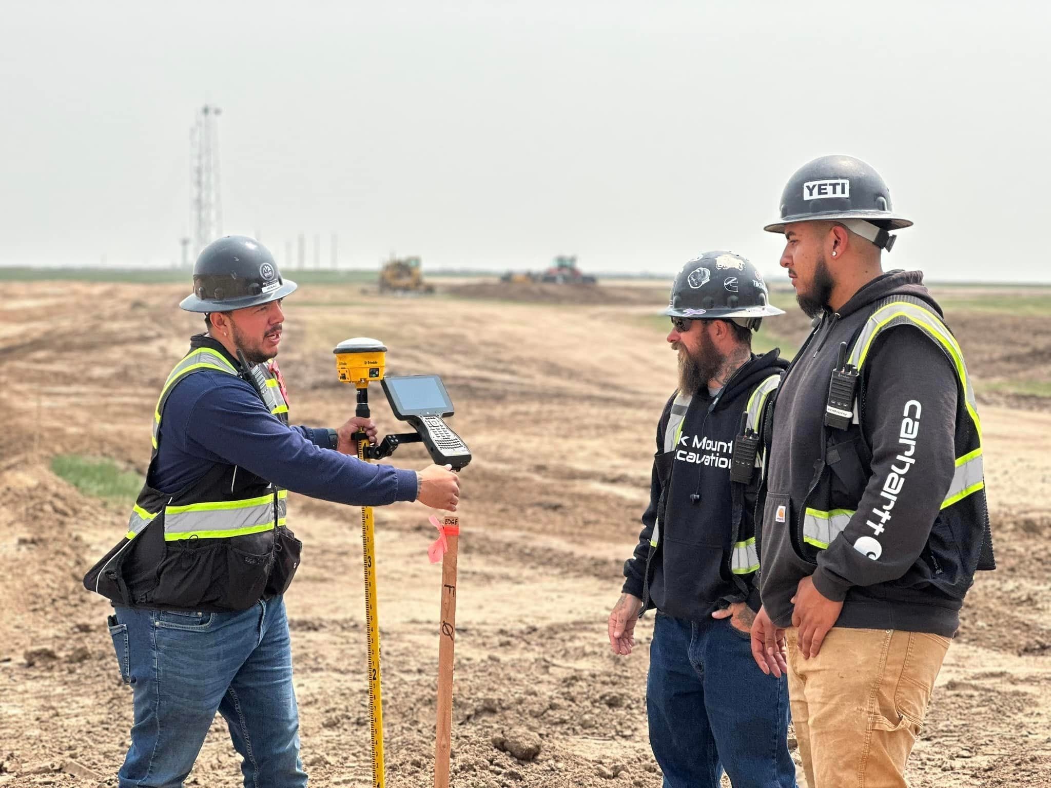 A BME surveyor holding a GPS rover and data collector while two crew members look on at an open job site
